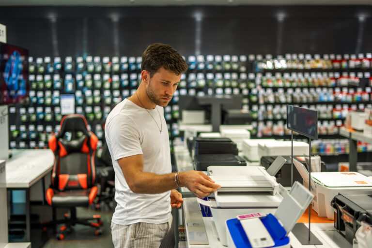 Side view of serious male customer choosing modern printer while standing at counter in electronics shop